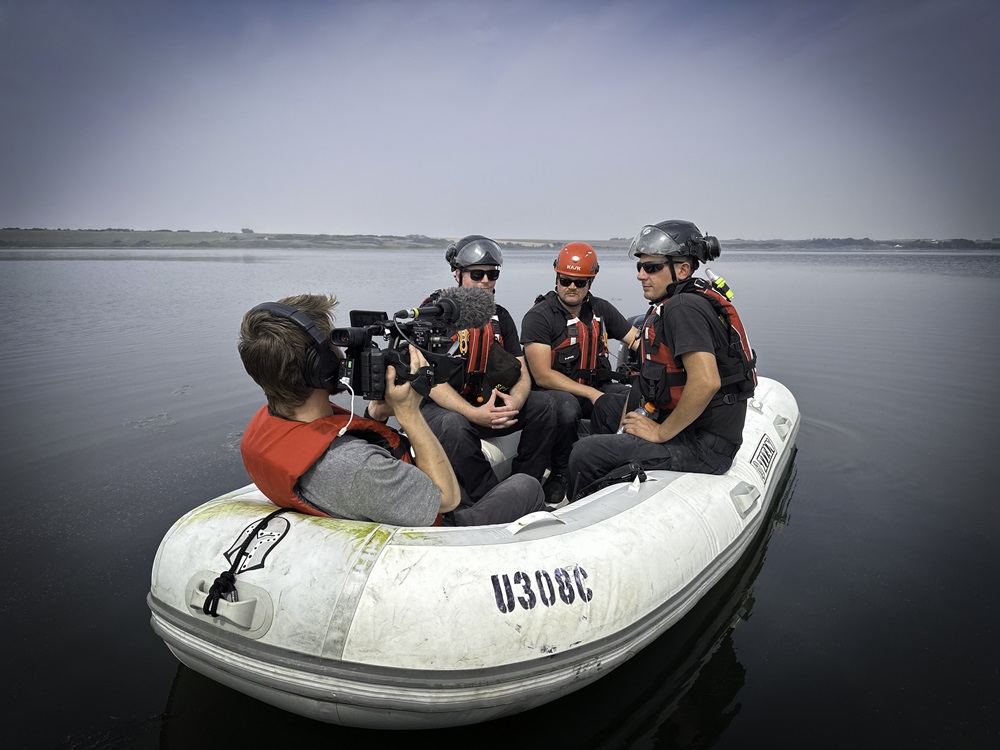 A camera man films three men in a boat on a lake